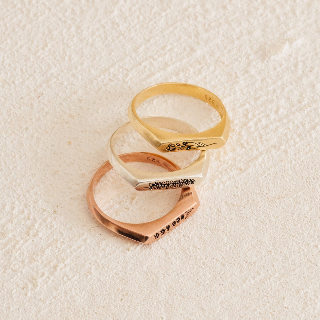 The Thin Flower Signet Ring, shown in gold, silver, and rose gold on a cream surface, features engraved floral details—making it the ideal personalized pinky ring for botanical jewelry enthusiasts.