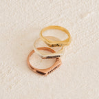 The Thin Flower Signet Ring, shown in gold, silver, and rose gold on a cream surface, features engraved floral details—making it the ideal personalized pinky ring for botanical jewelry enthusiasts.