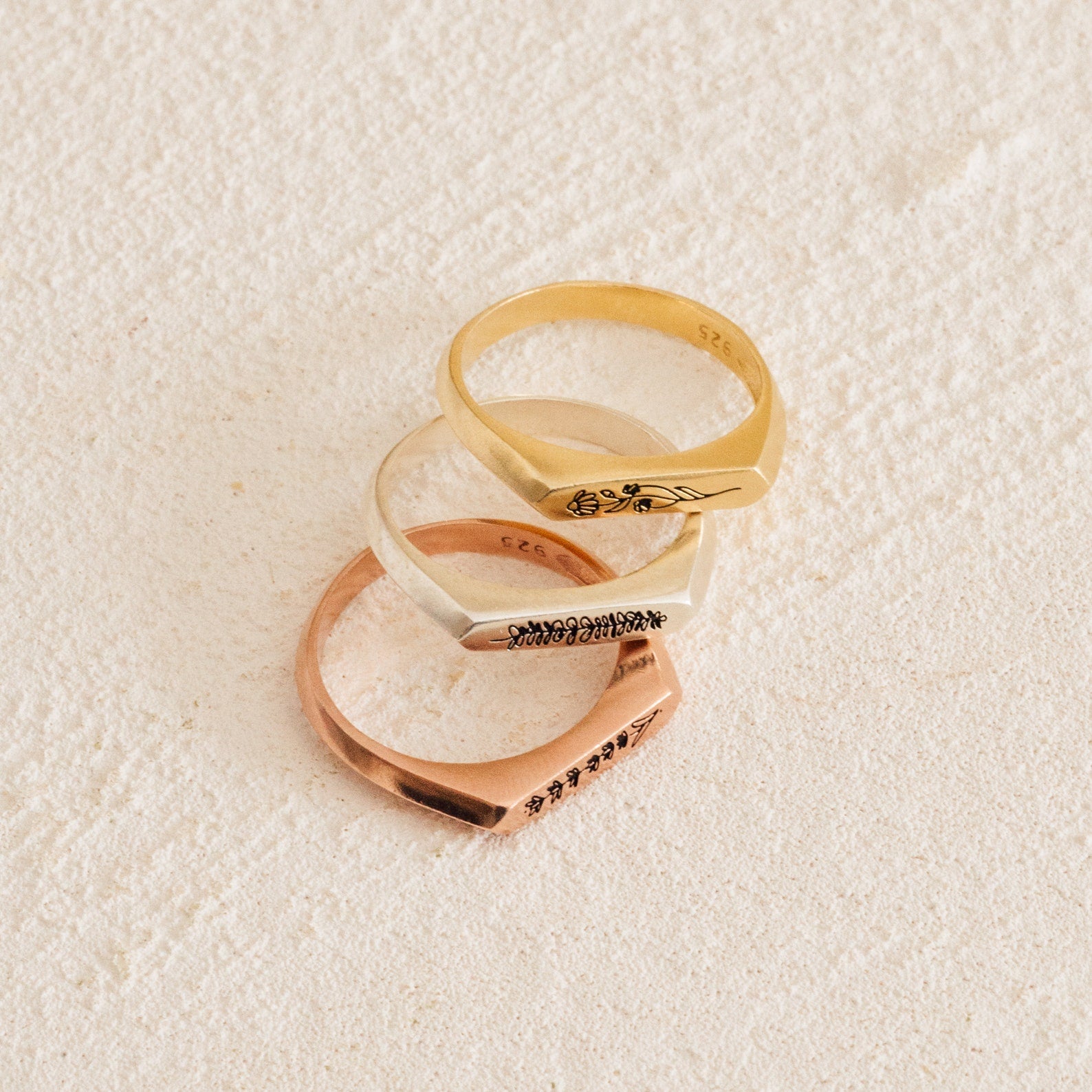 The Thin Flower Signet Ring, shown in gold, silver, and rose gold on a cream surface, features engraved floral details—making it the ideal personalized pinky ring for botanical jewelry enthusiasts.