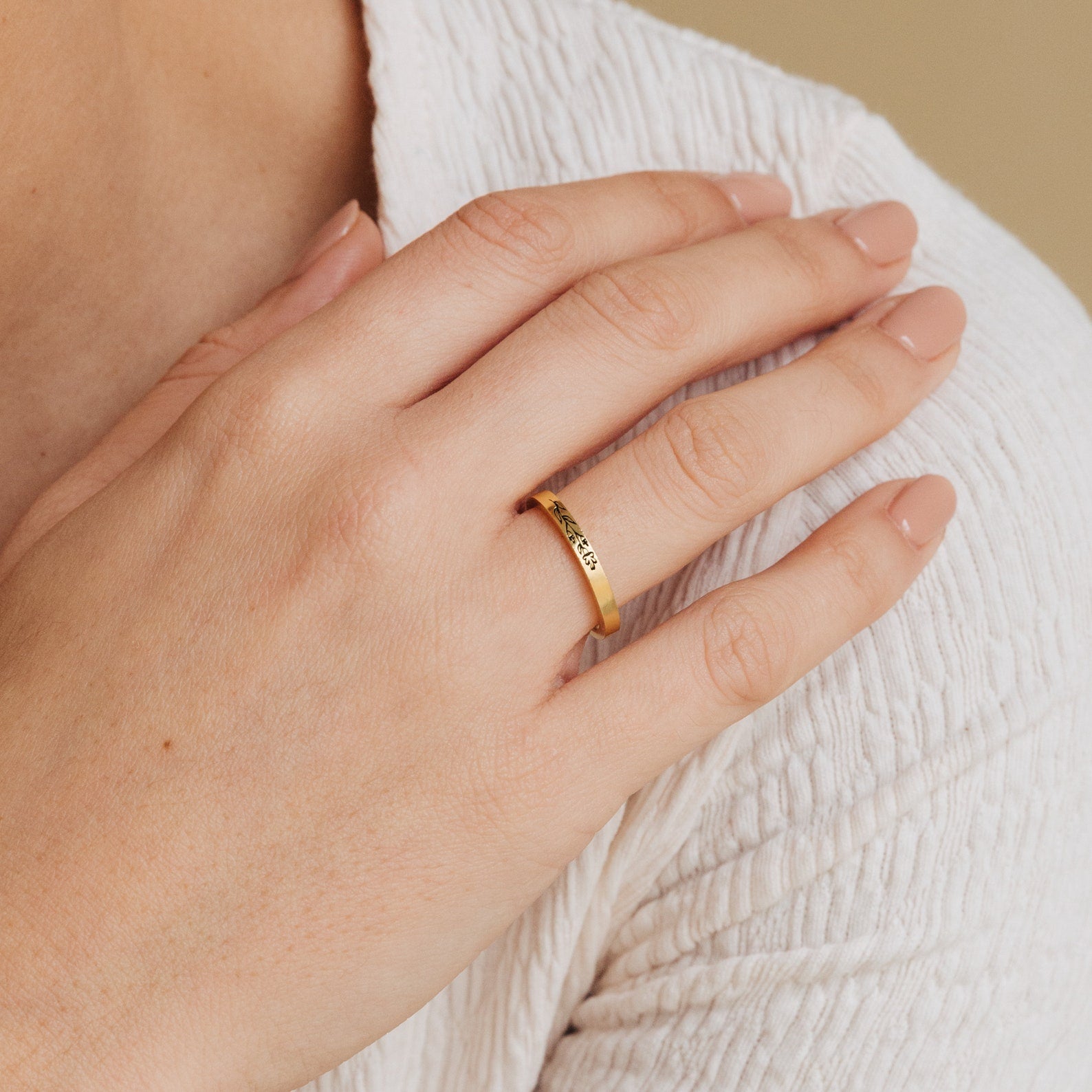 A hand with light pink nails wears the Stacking Flower Band, its gold botanical design gleaming as it rests on a textured white sleeve.