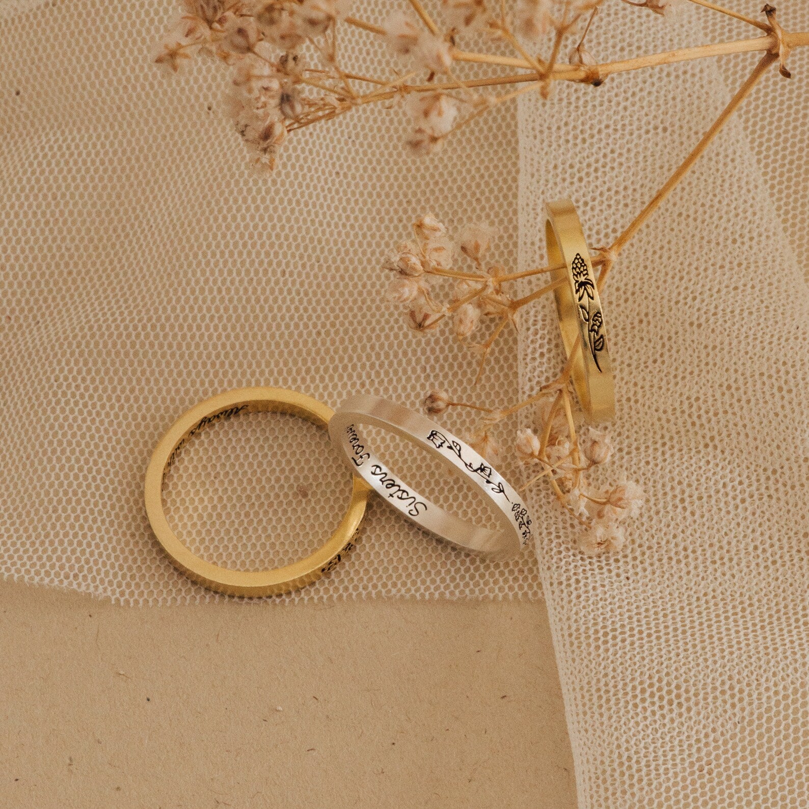 The Stacking Flower Band, alongside a slim gold band and an engraved silver ring, is displayed on tulle beside dried flowers, highlighting the botanical elegance of this jewelry piece.