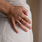 A hand with four gold rings, including a custom Flower Name Ring, rests against white fabric as the fingers gently curve.