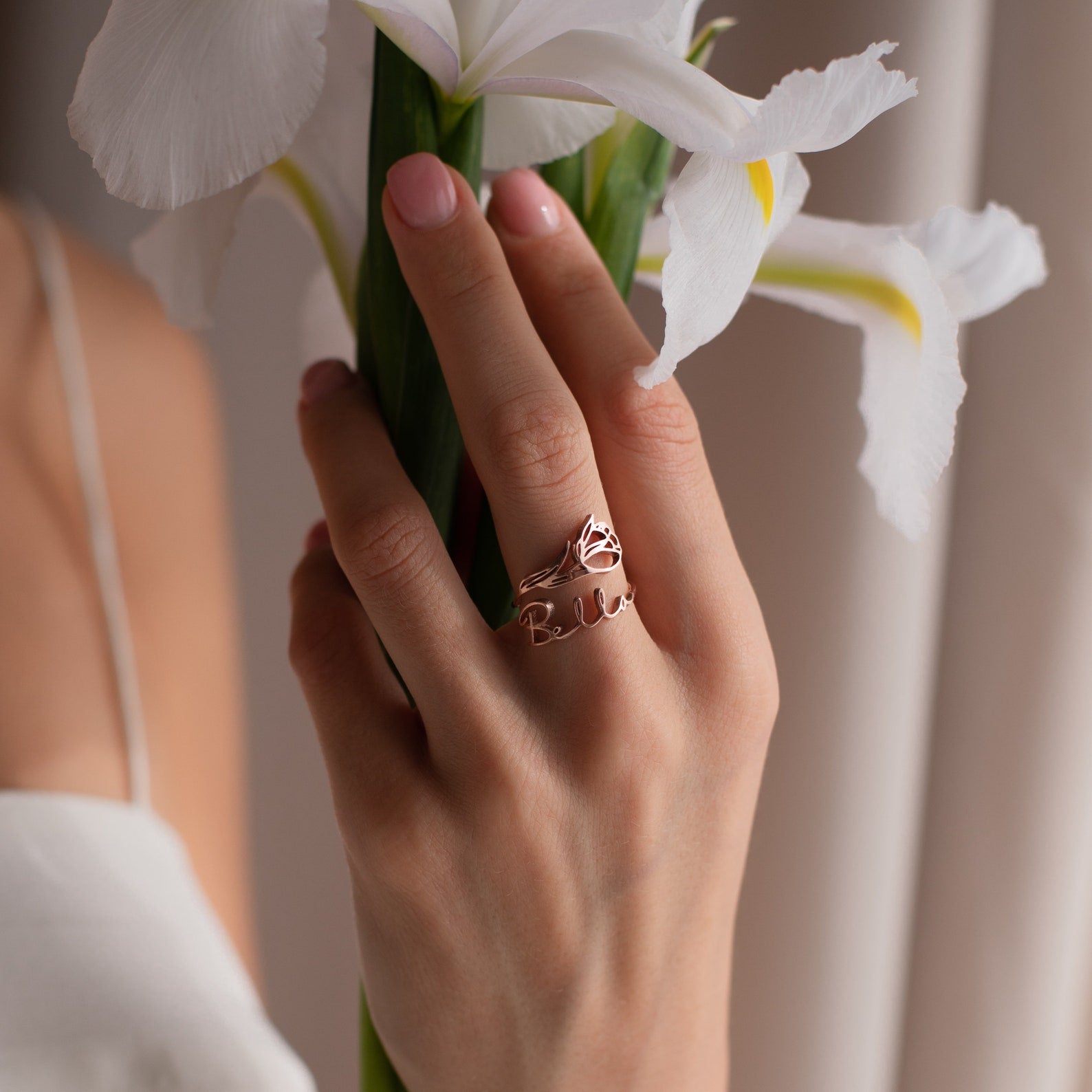 A hand wearing a rose gold Flower Name Ring holds a white iris; paired with a white top, this elegant ring makes an ideal gift for moms.