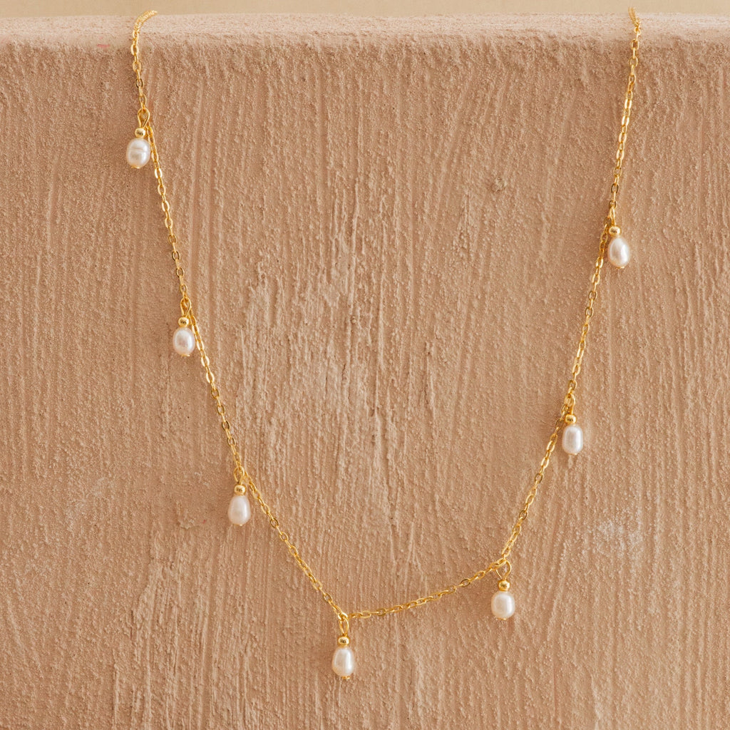 Delicate gold chain necklace with small dangling freshwater pearls, displayed against a textured beige surface.