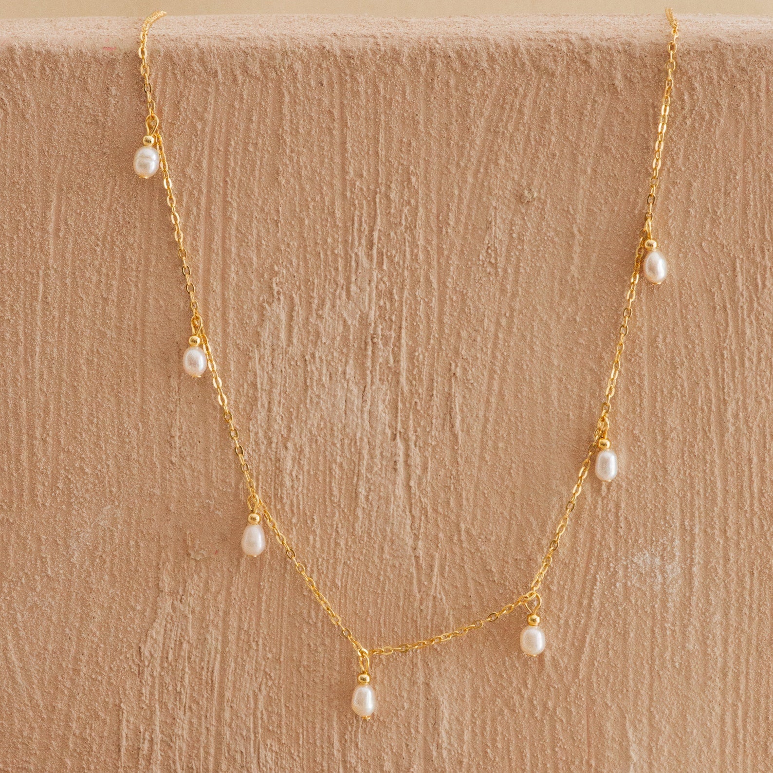 Delicate gold chain necklace with small dangling freshwater pearls, displayed against a textured beige surface.