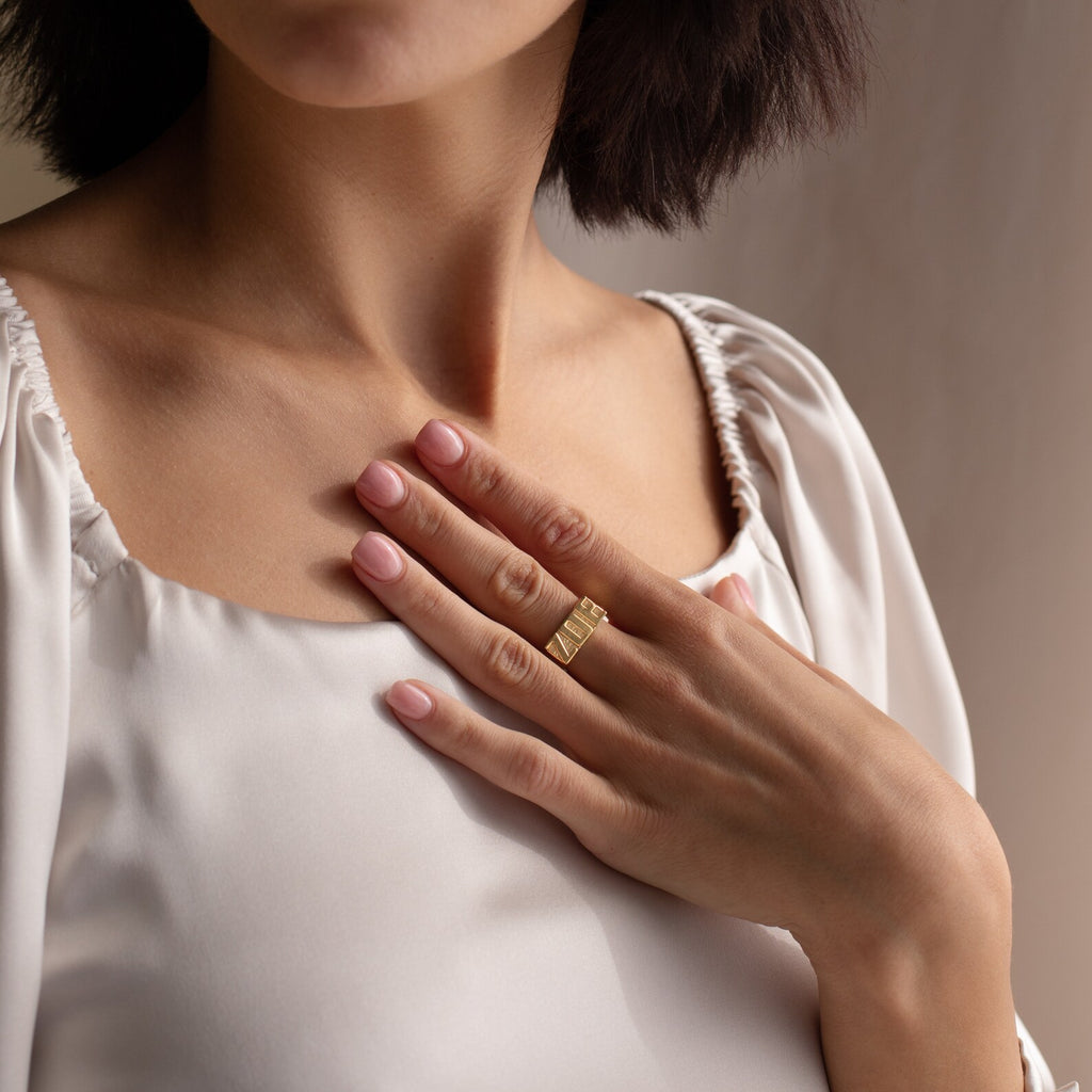 A woman touches her chest, wearing a white blouse and showcasing the Bold Name Ring in gold on her finger.