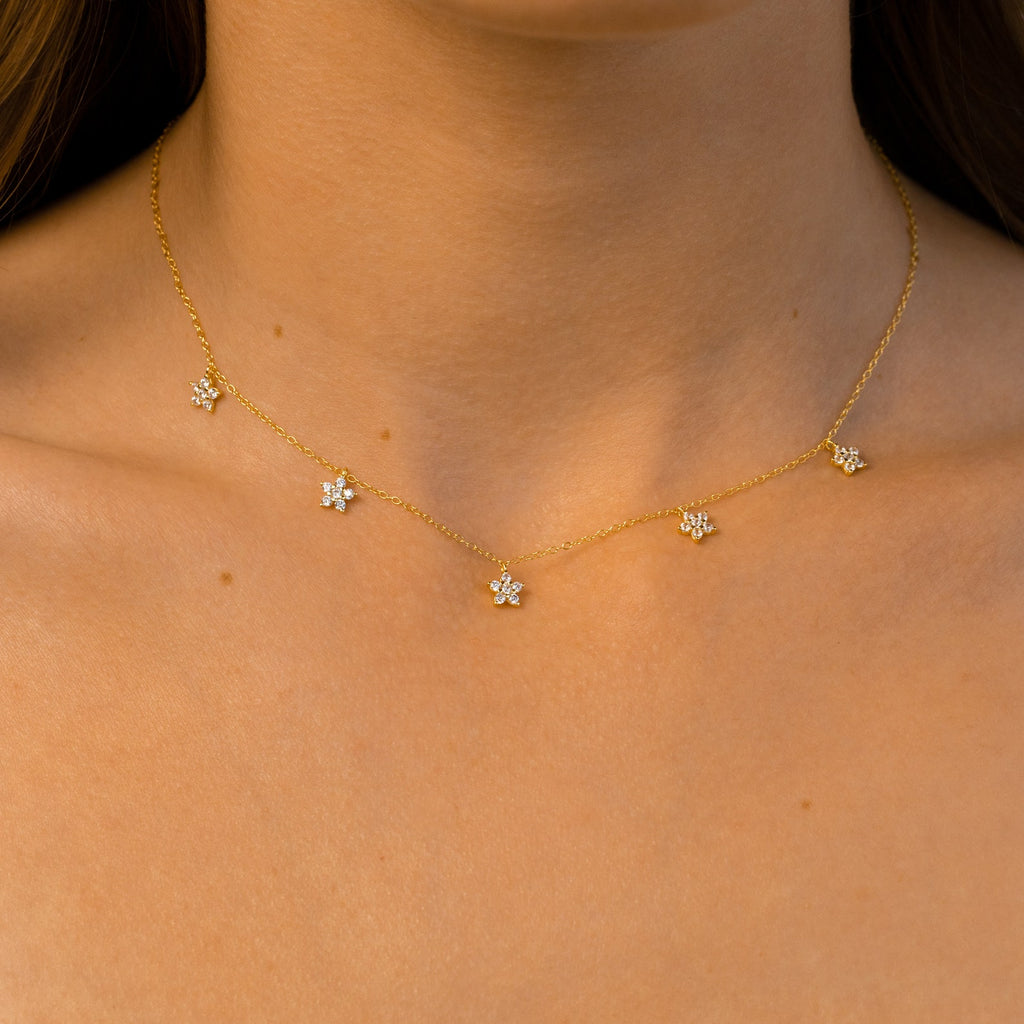 Close-up of a gold necklace with small flower-shaped crystal charms spaced along a fine chain, worn against bare skin in warm, natural lighting.
