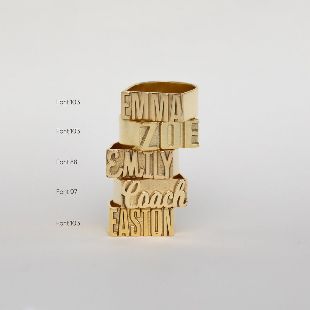 Five Bold Name Rings, each engraved with a unique name and font, are stacked vertically on a plain white background to create a striking statement set.