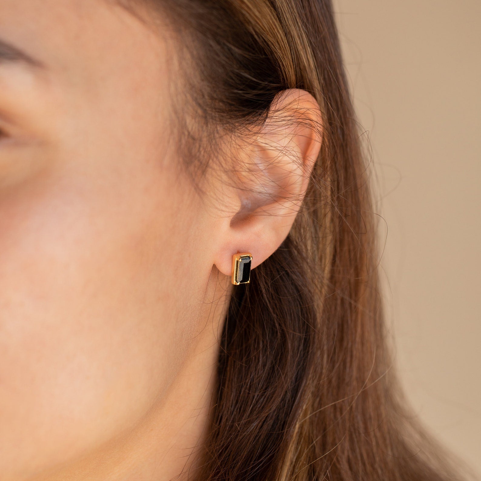 A close-up of a woman's ear wearing Rectangle Black Onyx Studs highlights the elegant black and gold earrings.