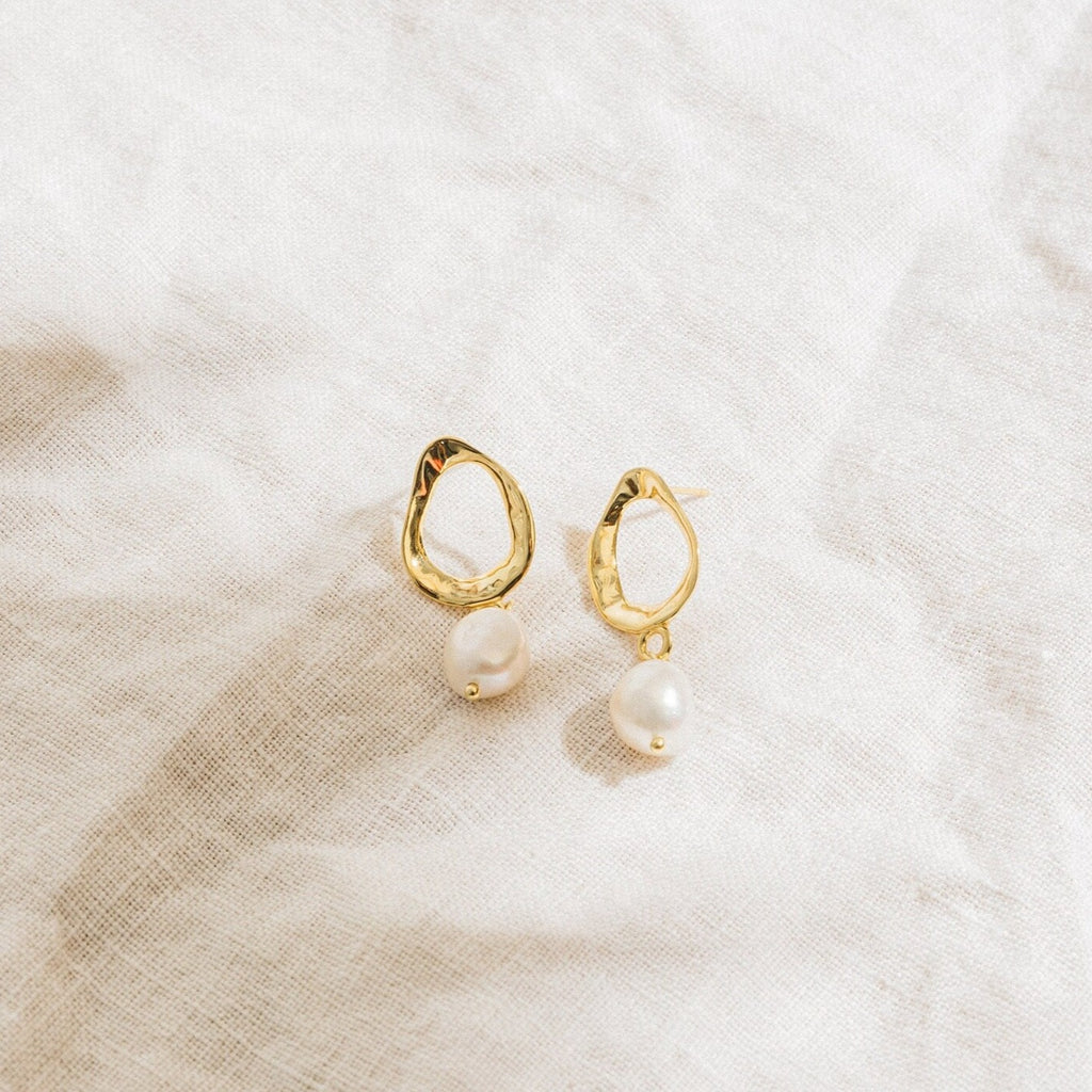 The Rebecca Pearl Drop Earrings feature gold, irregular oval shapes with freshwater pearl drops, displayed on a light, textured fabric background.