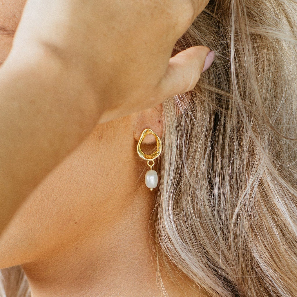 Close-up of a woman’s ear adorned with Rebecca Pearl Drop Earrings in gold, featuring a lustrous freshwater pearl, as her hand gently touches her hair.