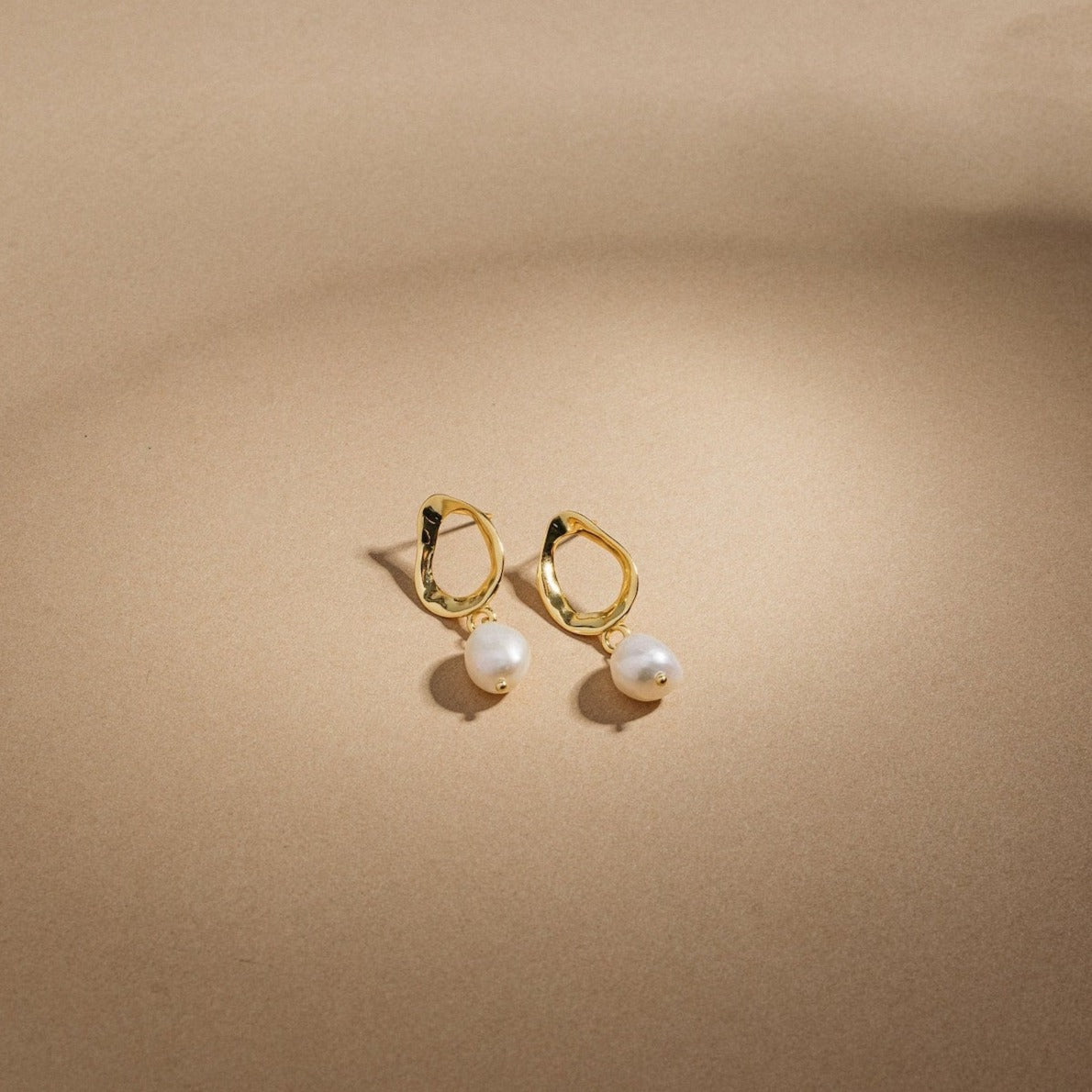 Rebecca Pearl Drop Earrings—gold hoops adorned with dangling freshwater pearls, shown on a beige background in soft light. These elegant earrings are the perfect accessory for any occasion.