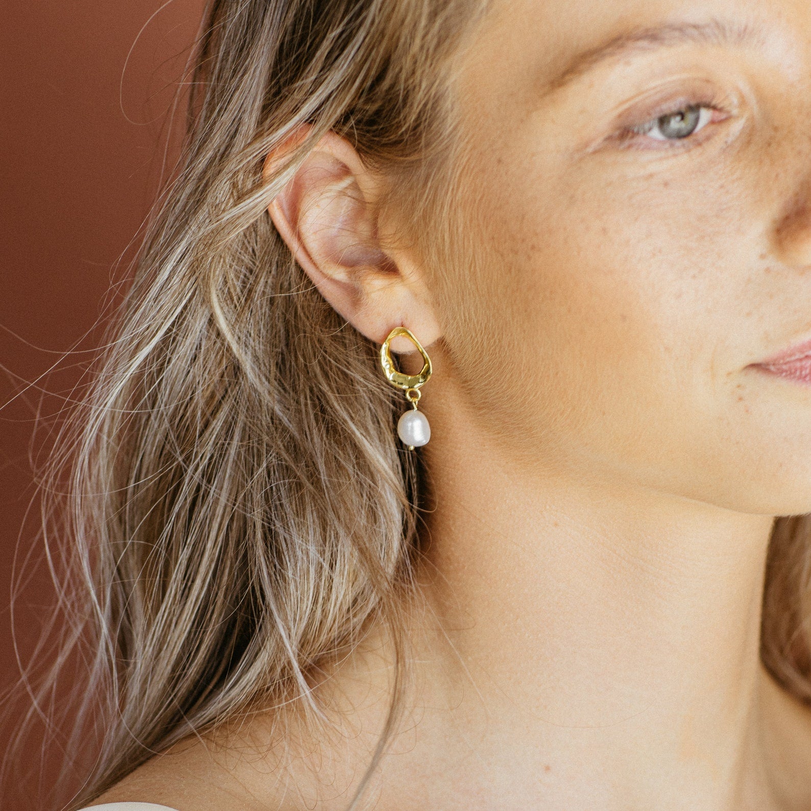 A woman with blonde hair wears Rebecca Pearl Drop Earrings—gold hoops featuring a shimmering freshwater pearl drop.