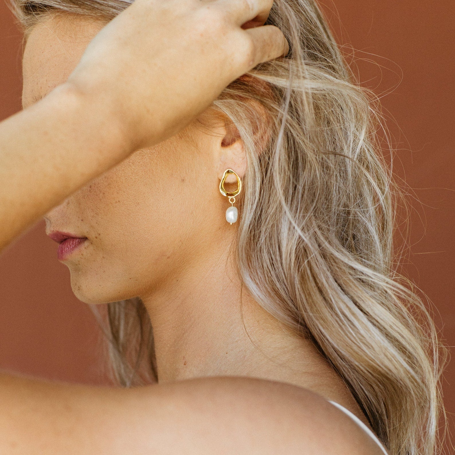 A blonde woman tucks her hair behind her ear, showcasing the elegant Rebecca Pearl Drop Earrings against a brown background.