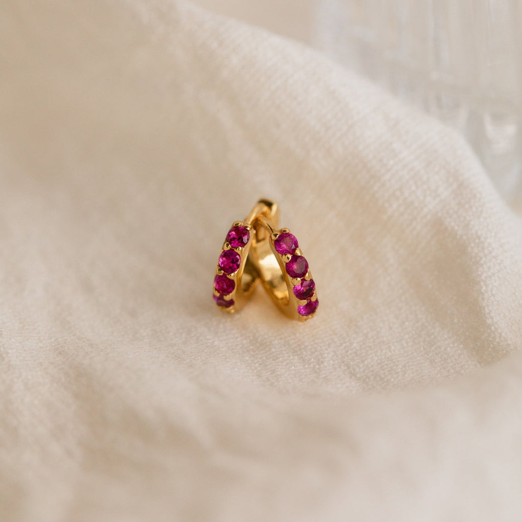 The Elise Gemstone Huggies Set features artisan-made earrings adorned with small pink stones, elegantly displayed on soft cream-colored fabric.