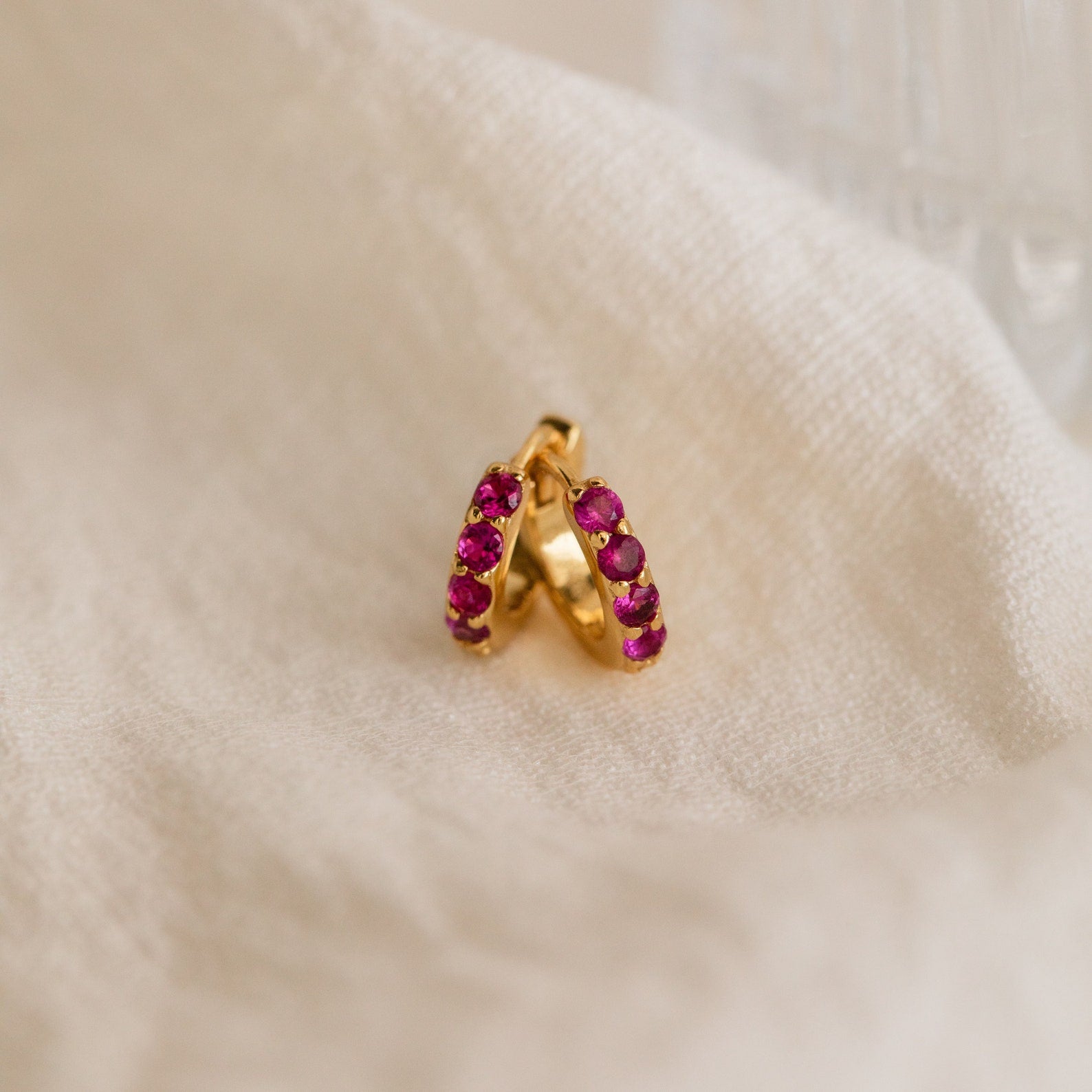The Elise Gemstone Huggies Set features artisan-made earrings adorned with small pink stones, elegantly displayed on soft cream-colored fabric.