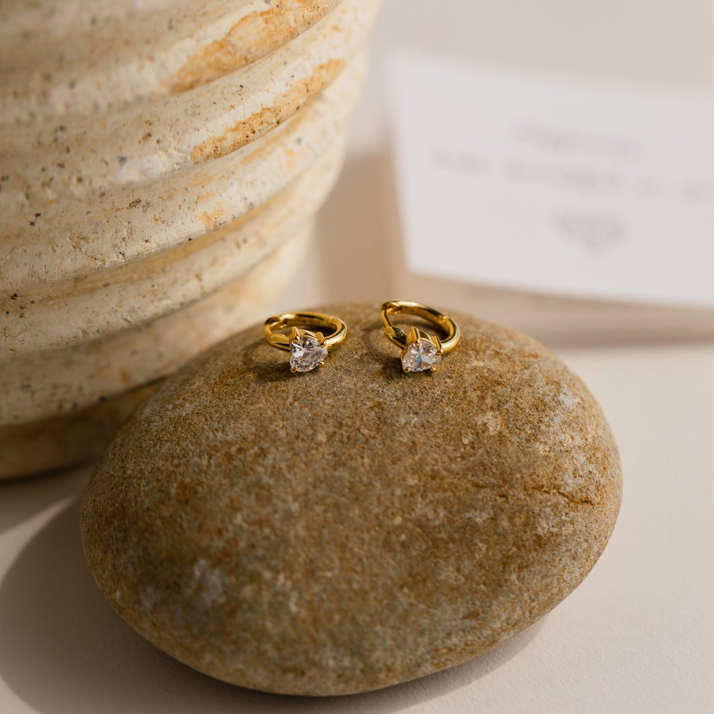 Diamond Heart Huggies with sparkling gemstones rest on a smooth stone beside a textured beige vase, making them an ideal gift for any occasion.