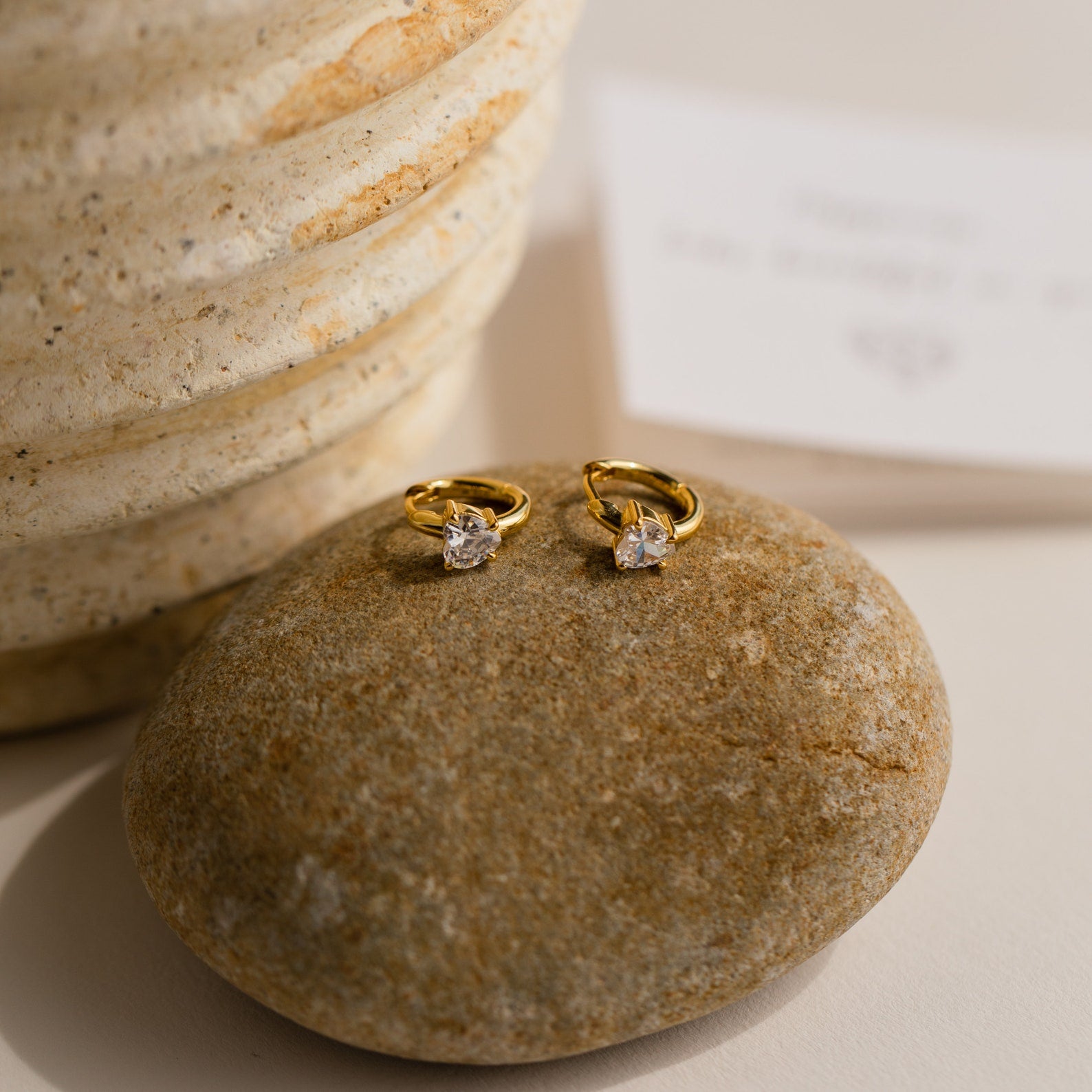 Diamond Heart Huggies with sparkling gemstones rest on a smooth stone beside a textured beige vase, making them an ideal gift for any occasion.