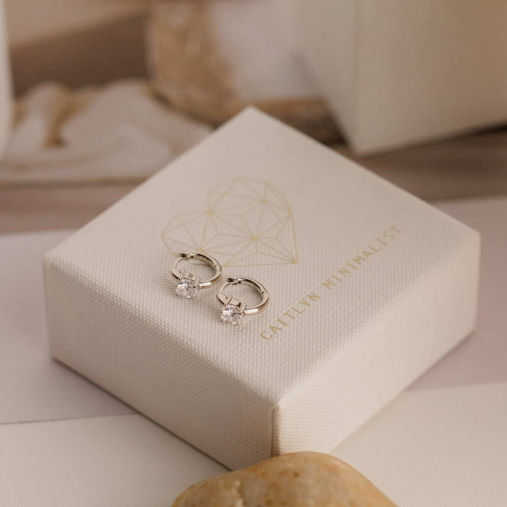 Diamond Heart Huggies with sparkling diamond accents rest on a white jewelry box adorned with a gold geometric heart—these dainty earrings are the perfect gift for someone special.