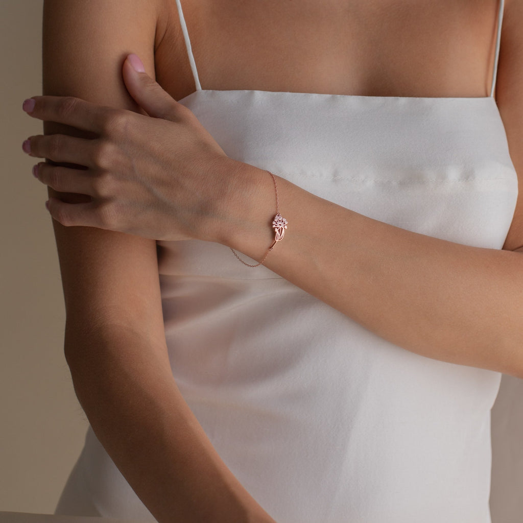 A woman in a white dress wears the Custom Flower Bracelet—a rose gold piece with a floral design on her left wrist, personalized with her unique birth flower, making it an ideal graduation gift.