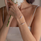 A woman in a white dress holds white flowers, wearing two delicate silver Flower Birthstone Bracelets on her wrist.