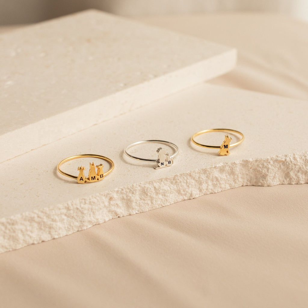 Three delicate Pet Initial Rings, each engraved with initials A, M, and B, are displayed on textured cream surfaces—a perfect personalized jewelry gift for pet lovers.