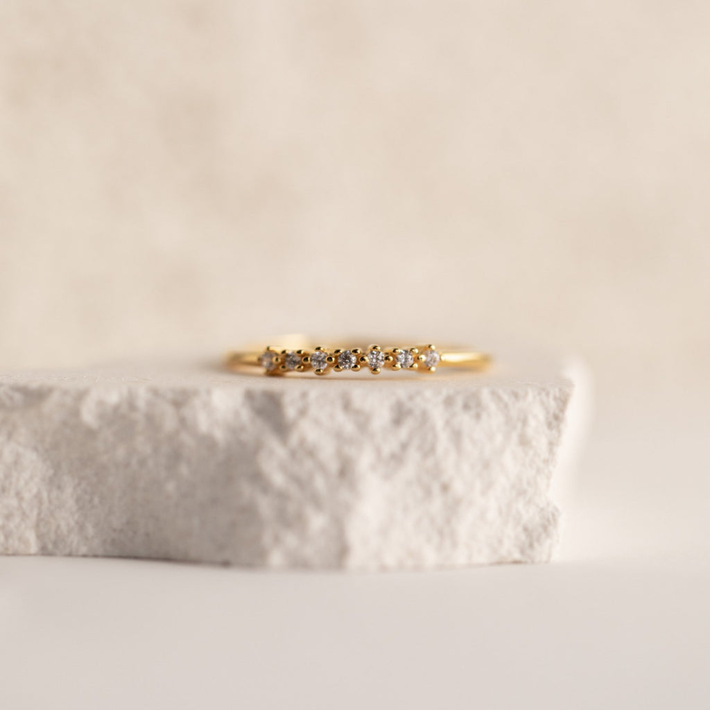 The Isabelle Diamond Ring, a delicate gold band adorned with small diamonds, is shown resting on a textured white stone surface.