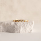The Isabelle Diamond Ring, a delicate gold band adorned with small diamonds, is shown resting on a textured white stone surface.