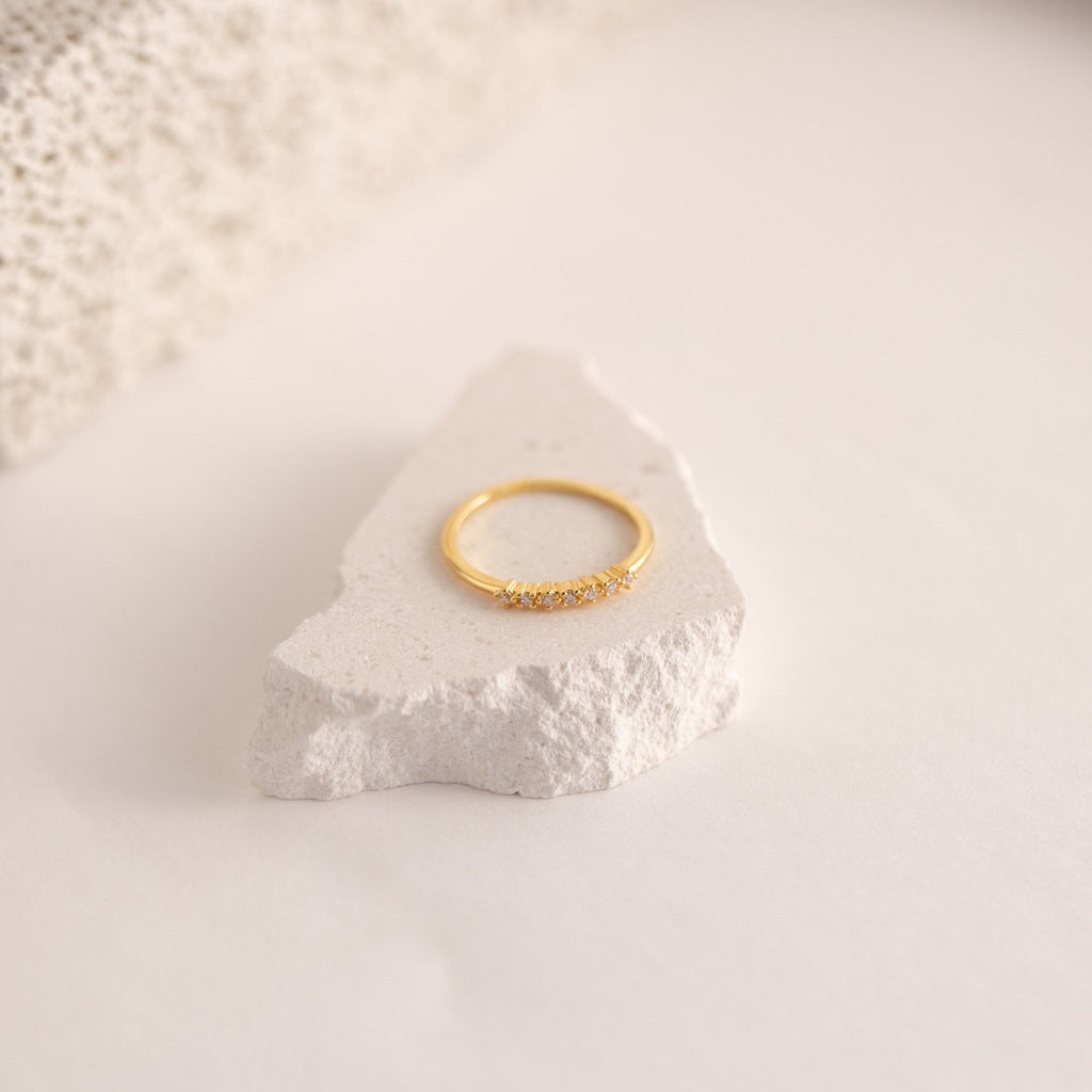 The Isabelle Diamond Ring, a delicate gold band accented with small diamonds, is showcased on a textured white stone set against a soft neutral backdrop.