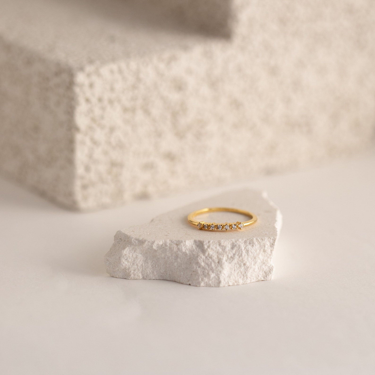 The Isabelle Diamond Ring, a delicate gold band with small diamonds, is shown on a white stone against a textured neutral background.