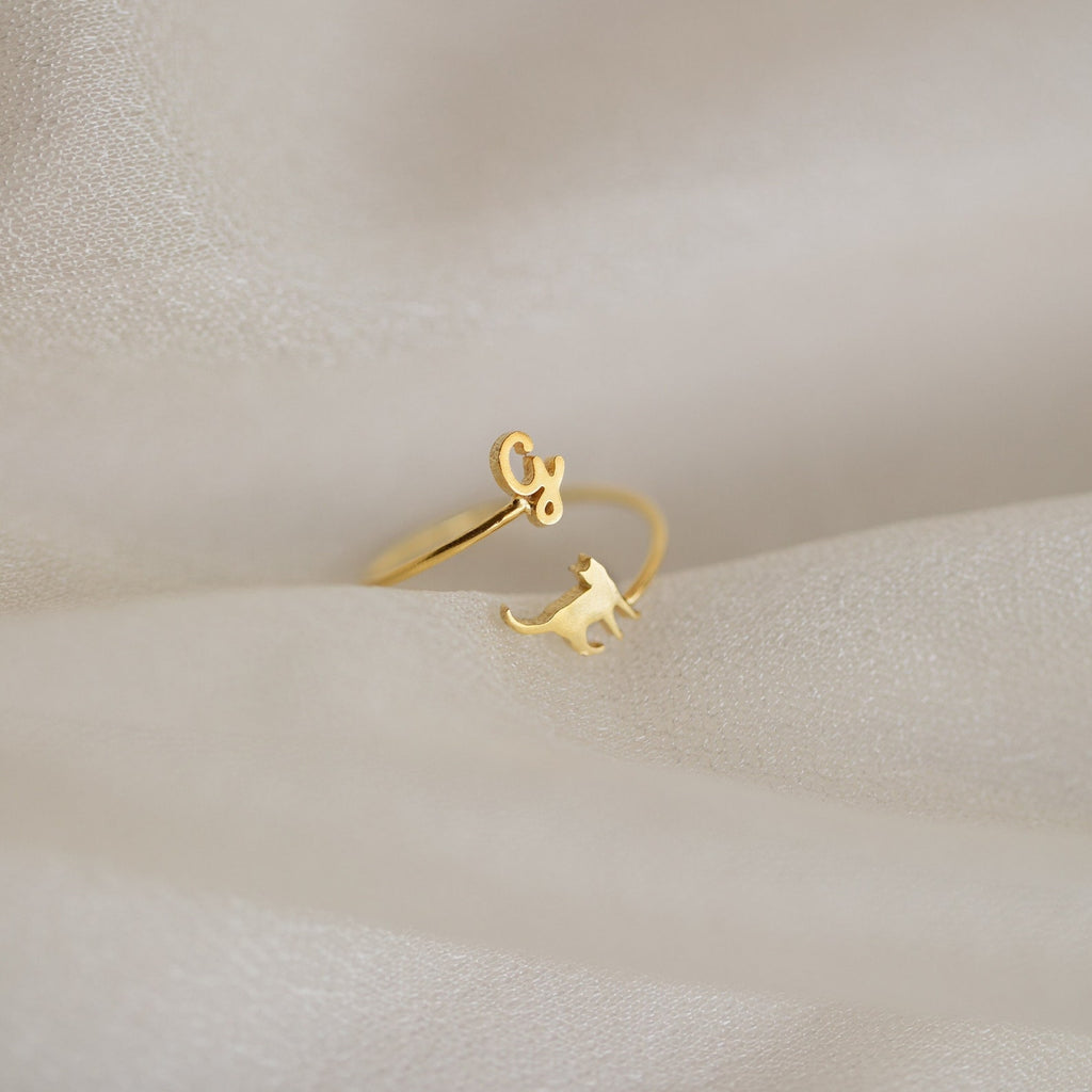 The Duo Pet Initial Wrap Ring showcases a cat and cursive "G" design, displayed on soft beige fabric—an ideal personalized jewelry piece for cat lovers.