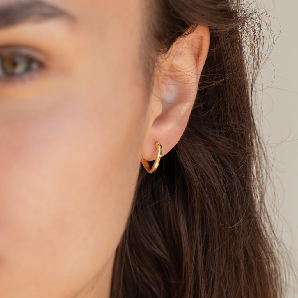 Close-up of a person’s ear wearing the McKenna Abstract Hoops—small gold earrings with an organic shape; brown hair partially visible.
