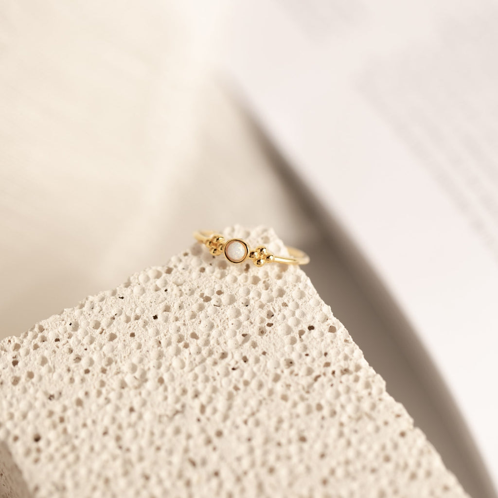 The Sophia Opal Ring is a vintage-inspired gold ring featuring a small white opal stone, showcased on a textured, light-colored surface.