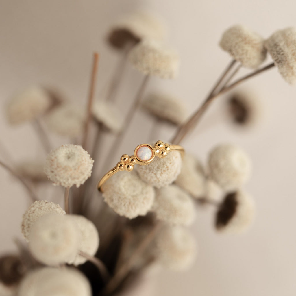 The Sophia Opal Ring, featuring a gold band and opal stone, rests on soft, fluffy white dried flowers against a neutral background.