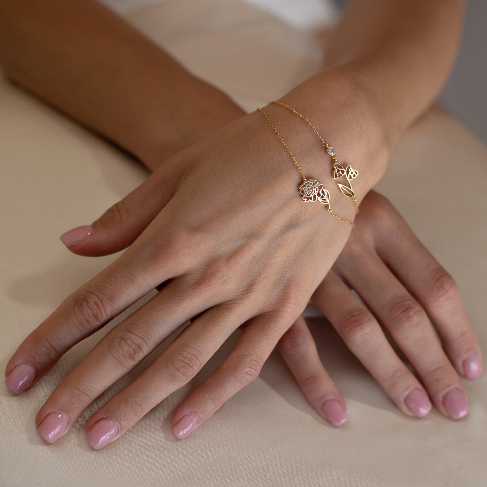 Two hands with light pink manicured nails wear delicate gold charm bracelets, including the Flower Birthstone Bracelet—an elegant and thoughtful birthday gift.