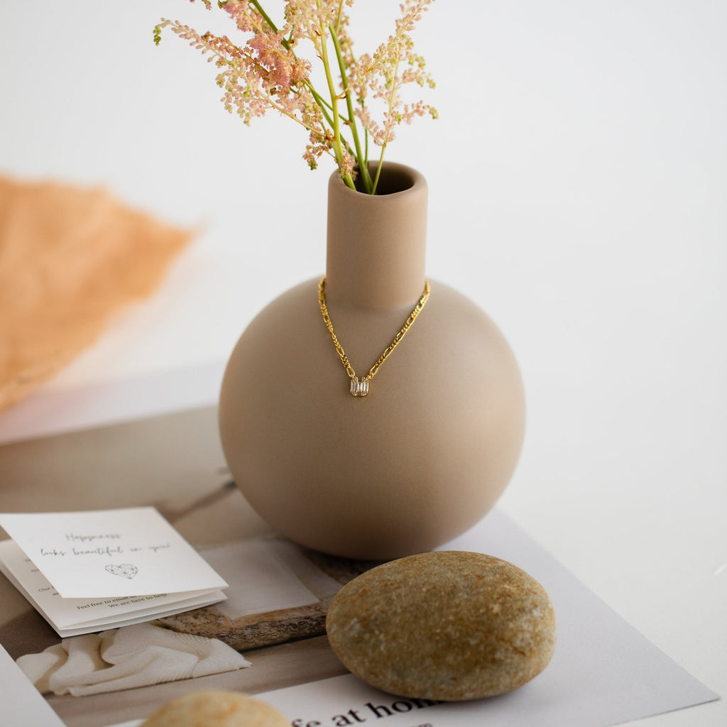 A Square Diamond Figaro Necklace drapes over a matte beige vase filled with pink flowers, placed atop a magazine with smooth stones beside it.