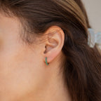 Close-up of a person’s ear wearing Ireland Emerald Huggies—lightweight small hoop earrings adorned with green gemstones.