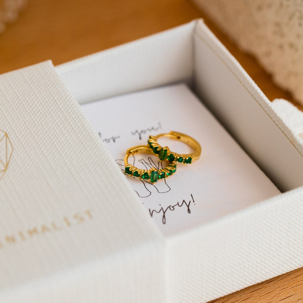 The Ireland Emerald Huggies, a pair of gold hoops with green stones, are displayed in a white jewelry box on a wooden surface.