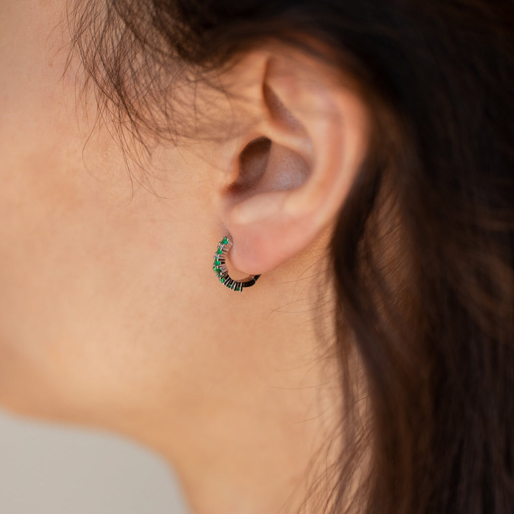 A close-up of a woman's ear wearing the Ireland Emerald Huggies—lightweight small hoop earrings adorned with green and black stones for an elegant look.