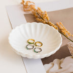 Four lightweight huggie hoop earrings—gold and silver, including Ireland Emerald Huggies—rest in a scalloped white dish with dried flowers, perfect for any look.