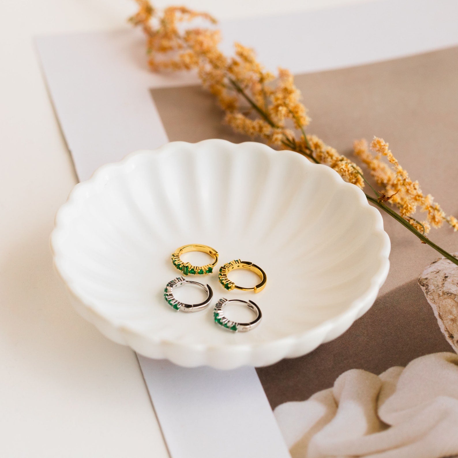 Four lightweight huggie hoop earrings—gold and silver, including Ireland Emerald Huggies—rest in a scalloped white dish with dried flowers, perfect for any look.