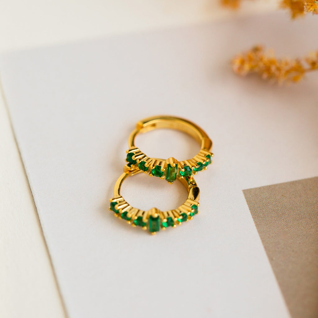 Our Ireland Emerald Huggies are lightweight gold hoop earrings with green gemstones, shown on a white and beige background accented by dried flowers.