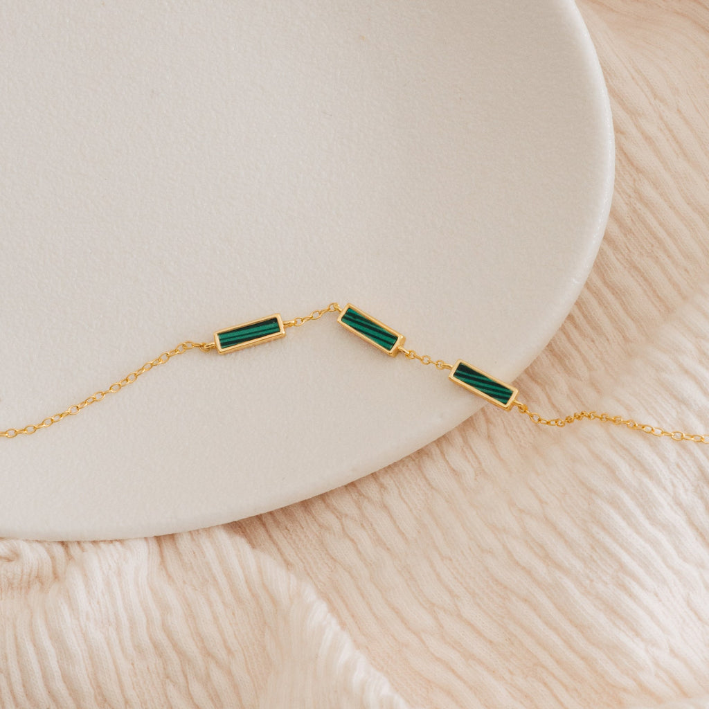 The Malachite Bar Bracelet is a gold dainty chain featuring three rectangular green malachite stones, handcrafted for a boho vibe and shown on a white plate with a soft beige fabric background.