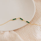 The Malachite Bar Bracelet is a gold dainty chain featuring three rectangular green malachite stones, handcrafted for a boho vibe and shown on a white plate with a soft beige fabric background.