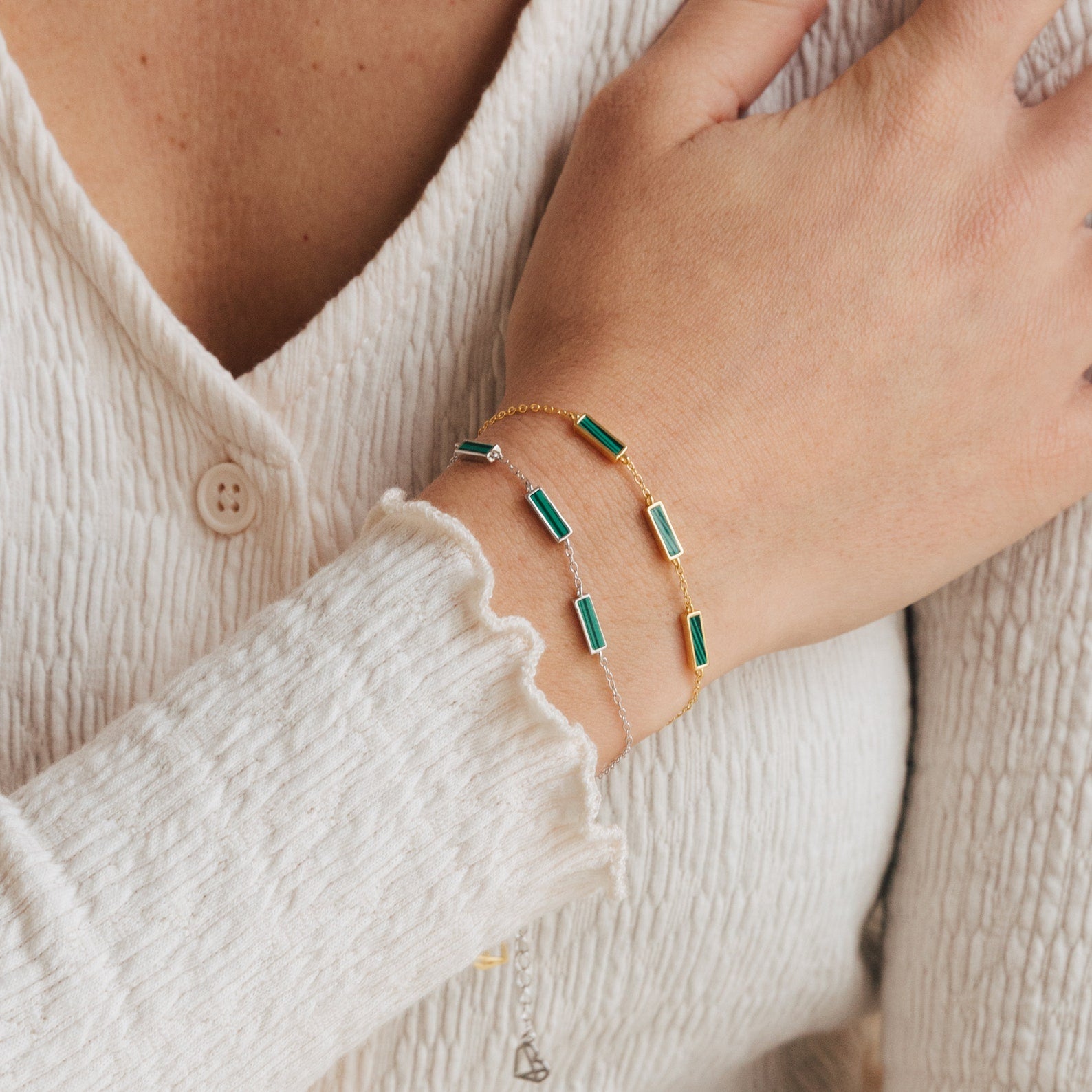A hand wearing the Malachite Bar Bracelet, featuring green rectangular stones and delicate chain details, rests on a textured white sleeve.