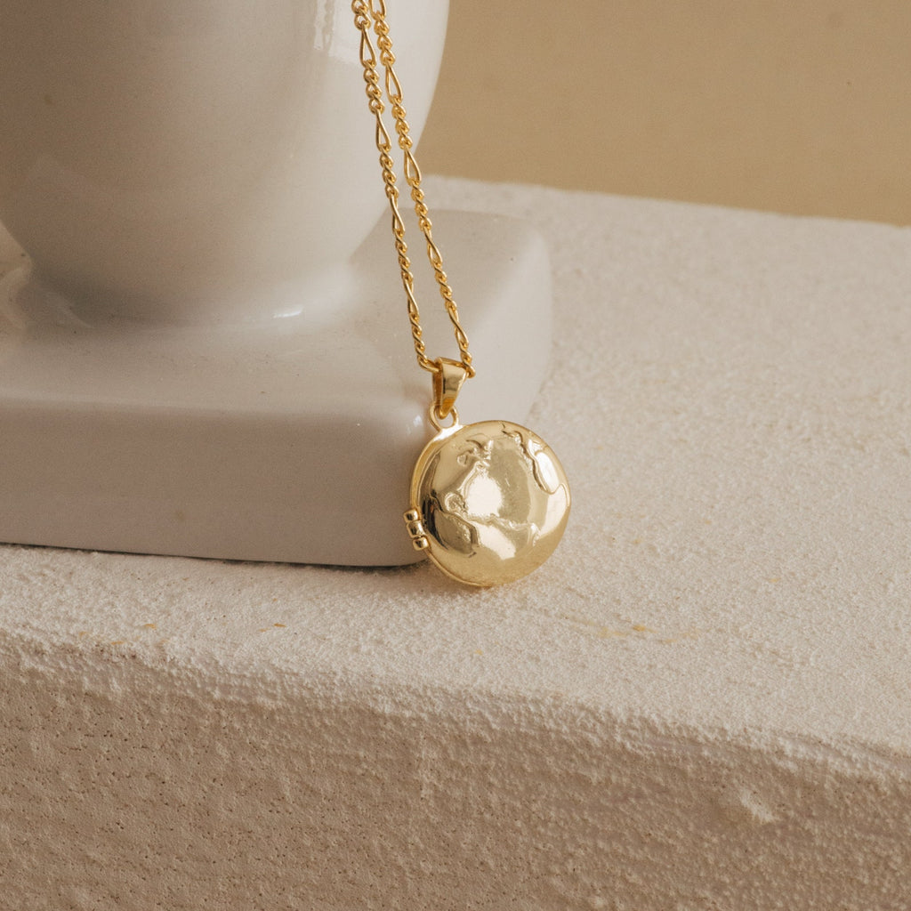 The Earth Locket Necklace features a globe design and is displayed against a white ceramic vase on a textured surface—an ideal travel keepsake for those who love nature-inspired jewelry.