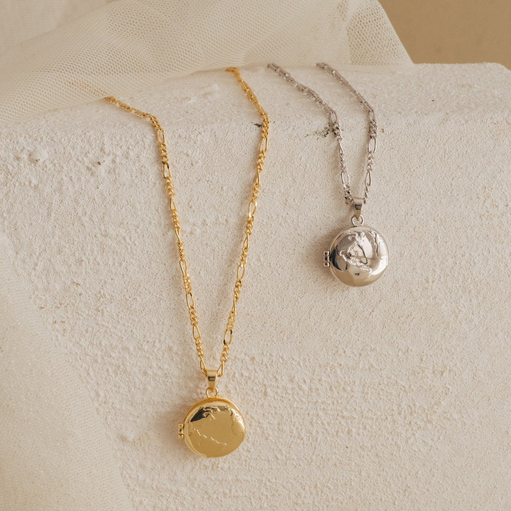 Two Earth Locket Necklaces—one gold, one silver—are displayed on a white stone surface with fabric. Each necklace is a textured keepsake, perfect as nature-inspired travel jewelry.