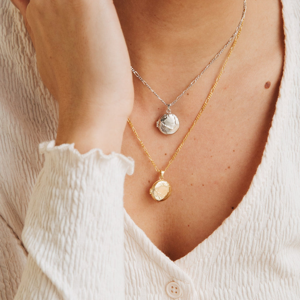 A person in a cream textured top and two coin necklaces, one gold and one silver, completes their look with the Earth Locket Necklace—a stunning piece of nature jewelry that's an ideal travel keepsake.