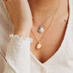 A person in a cream textured top and two coin necklaces, one gold and one silver, completes their look with the Earth Locket Necklace—a stunning piece of nature jewelry that's an ideal travel keepsake.