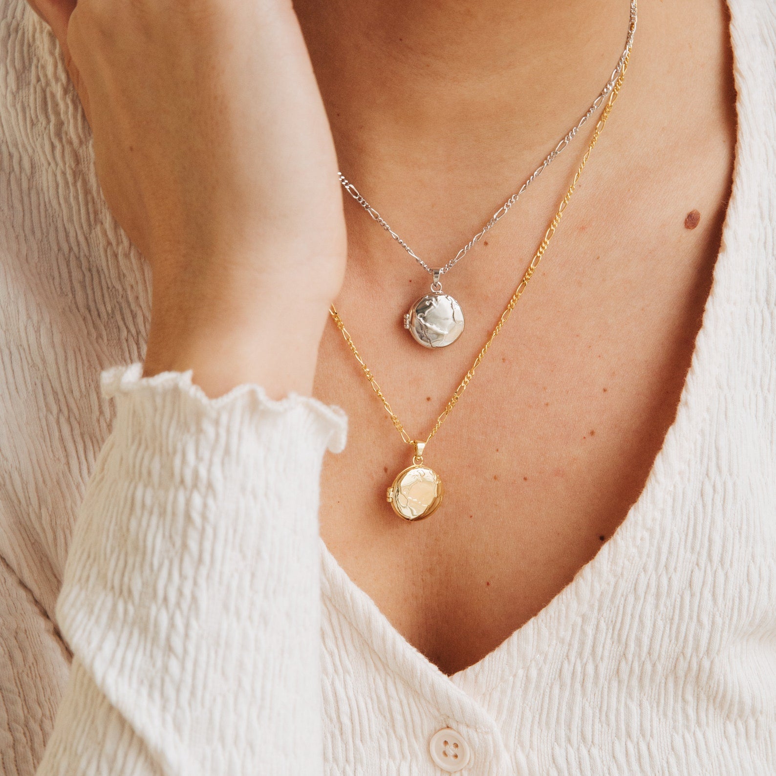 A person in a cream textured top and two coin necklaces, one gold and one silver, completes their look with the Earth Locket Necklace—a stunning piece of nature jewelry that's an ideal travel keepsake.